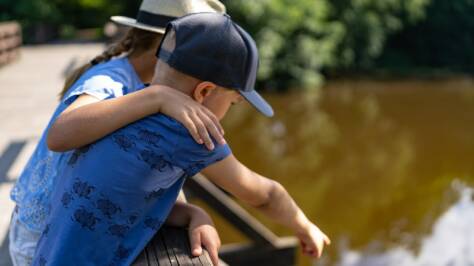 En flicka och en pojke står på en bro och håller om varandra medan de kollar ner i ån.